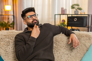 Indian man at home sits on sofa and gazes out at side with dreamy nostalgic expression on face....