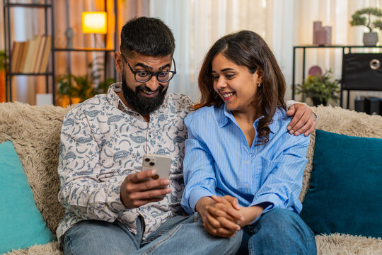 Indian couple man and woman sitting on home sofa, using smartphone for food order, joking and laughing while debating dinner. Young family enjoying playful moment with smiles and fun emotions indoors. - Powered by Adobe