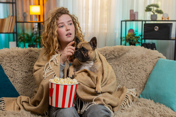 Young happy Caucasian redhead woman watches television with pug dog sits on home sofa, both wrapped...
