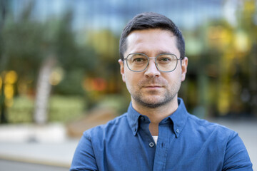 A man wearing glasses and a blue shirt looks directly at the camera with a serious expression,...