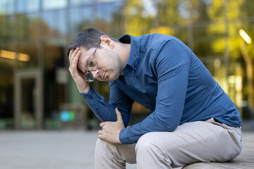 A man with glasses sits outside, looking down with a hand on his head, appearing stressed and...