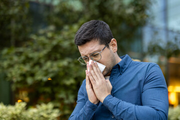 A man in glasses outside, blowing his nose with a tissue, suggesting allergies or illness.