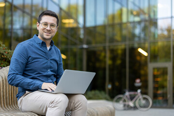 A smiling man with glasses uses a laptop outdoors, sitting on a bench in front of a modern...