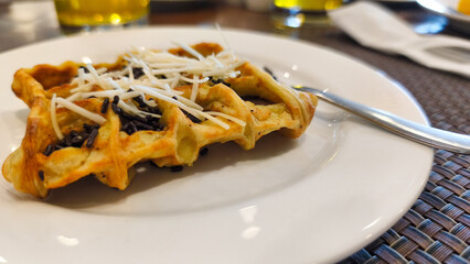 Close up of a golden brown waffle topped with grated cheese and chocolate sprinkles, served on a white plate with a fork beside it.