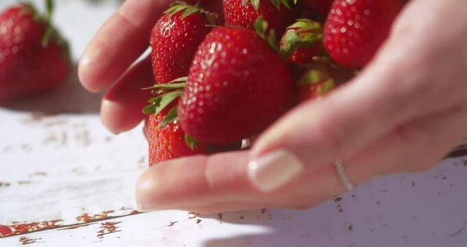 A woman's hand gently picks up a handful of vibrant strawberries and lets them fall slowly onto a rustic white table. Sunlight enhances the juicy texture in this soft, slow-motion kitchen scene.