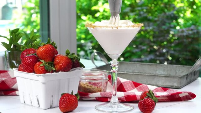 Strawberry Shortcake Martini cocktail with whipped cream, graham cracker crumbs and a fresh red strawberry. Piping the whipped cream on top. A summer time berry drink with red gingham napkin.