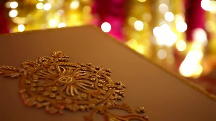 An intricately decorated red and gold Indian wedding invitation box lies open, with vibrant pink and yellow floral garlands blurred in the background. Concept of Indian wedding traditions