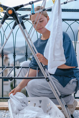 Young woman drying laundry on balcony during slow morning at home. Real moment, Gen Z lifestyle,...