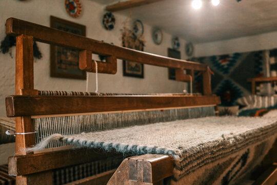 Handcrafted wooden loom with wool fabric partially woven, situated in cozy rustic interior with folk patterns on walls. Traditional textile production in warm artisan environment