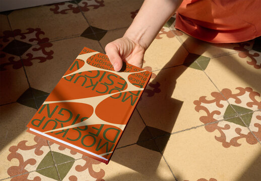 Woman holding a blank back cover notebook above a colorful, patterned tile floor. PSD template mockup