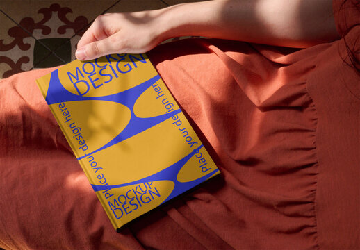 mockup of a blank notebook cover rests on woman's lap, casting a shadow on a warm surface