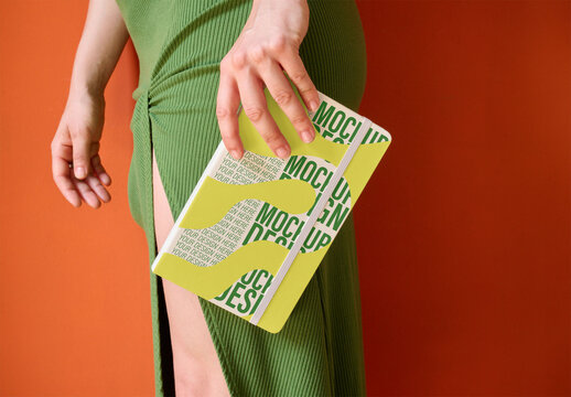 A stylish notebook mockup is being held by anonymous woman dressed in a green outfit against a vivid orange wall. PSD template mockup.