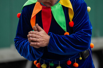 clown performer in colorful velvet costume with pom-poms holding hands together, festive jester...