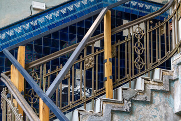 Fototapeta premium Damaged staircase with decorative wrought iron railing and blue tiled wall. Temporary structural supports made of wood and metal in an abandoned or renovated building.