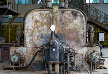 Vintage industrial generator with valves, pipes, and a pressure gauge, covered in rust and patina....