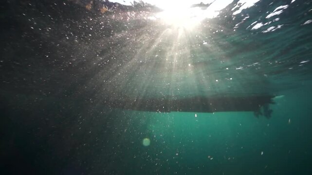 Sunlight breaks through the surface, illuminating particulate matter swirling below. A boat's hull and propeller appear silhouetted in the depths of the Arctic Barents Sea.