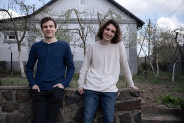 Two friends enjoy a sunny afternoon by a rustic stone wall in a serene garden setting, sharing...