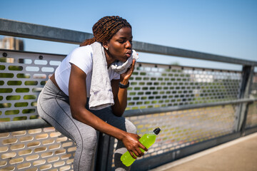 Portrait of exhausted African woman in sports clothing wiping sweat with towel, drinking energy...