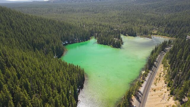 Drone descending on emerald green Devils lake with cars driving on Cascade Lakes highway in Central Oregon. Snow remaining on the shore of the lake.