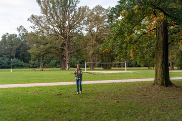 Fototapeta premium Young boy in plaid shirt and glasses holding long wooden stick standing on green grass in autumn park with trees and volleyball court, outdoor childhood adventure and nature exploration concept. 