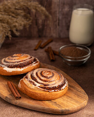 Two tasty homemade cinnamon rolls bun with sugar icing on wooden cutting board on dark brown table...