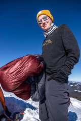 Smiling man holding a down sleeping bag looks at the camera outside orange tent on snowy mountain
