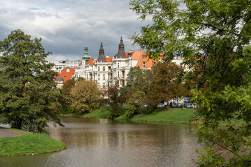 Obraz premium Historic European city architecture with red tile roofs and towers reflected in calm river water, framed by green trees under cloudy sky. Scenic urban landscape and cultural heritage concept. 