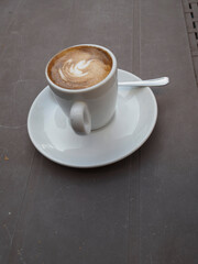 Ceramic cappuccino cup with intricate latte art pattern sitting on saucer near silver teaspoon, positioned on rustic wooden cafe table in Cremona, Lombardy