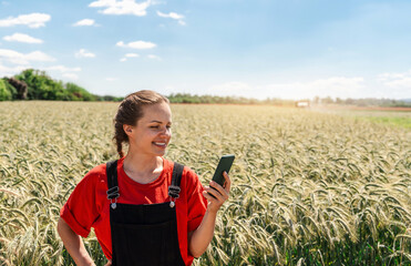 Female farmer checking her smart phone while standing near cereal field in mid season. Mobile...