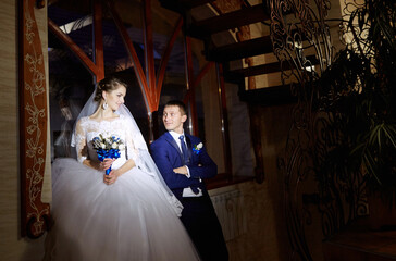 A bride and groom stand in front of a window, with the bride holding a bouquet