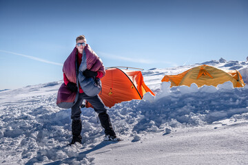 Man wrapped in a down sleeping bag stands on a snowy mountain near two orange tents. Sunny winter...