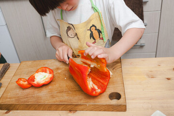 Child slicing red bell pepper on a wooden cutting board at home