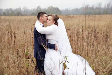 A bride and groom are standing in a field, with the bride wearing a white dress