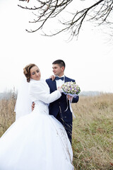 A bride and groom are standing in a field, with the bride holding a bouquet