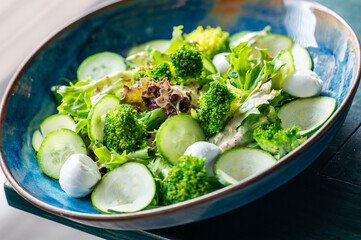 Fresh vegetable salad with broccoli, cucumber slices, mixed greens, and mozzarella balls in a blue bowl. Healthy, vibrant meal ideal for diet and wellness concepts.