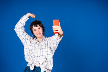 Young woman taking a selfie with smartphone and raising one arm on blue background