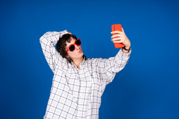 Young person taking a selfie with red smartphone and sunglasses on blue background