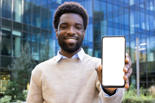 Smiling man holding up a phone with a blank screen in front of a modern glass building. The photo is taken in daylight.