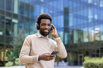 Smiling man wearing headphones holds a phone in front of a modern building with a glass facade.