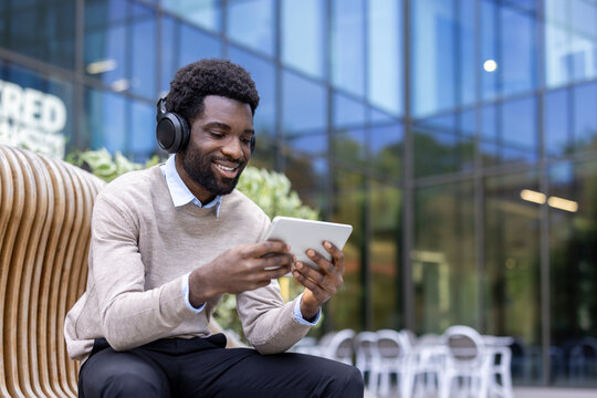 An african american man with headphones smiles as he looks at a tablet outdoors in front of a building.