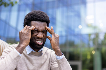A man experiencing a headache, pressing his fingers to his temples with eyes closed outdoors.