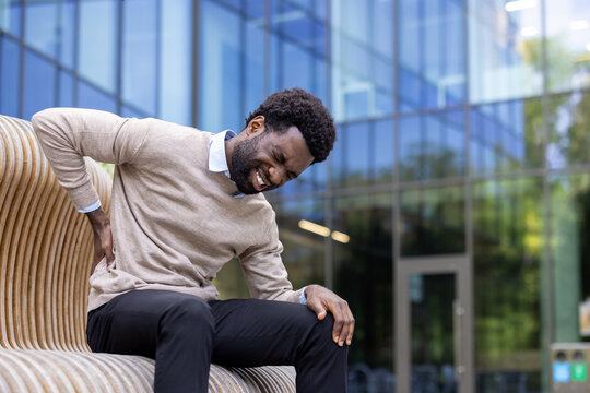 An african american man is experiencing back pain while sitting on a bench outside.