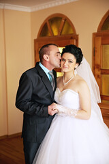 A bride and groom are kissing in front of a stone wall. The bride is wearing a white dress and the...