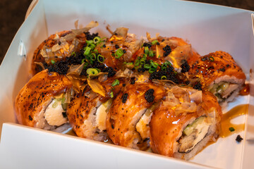 Close-up of assorted sushi rolls in a takeout box, topped with sauce, roe, and green onions, highlighting Japanese cuisine freshness.