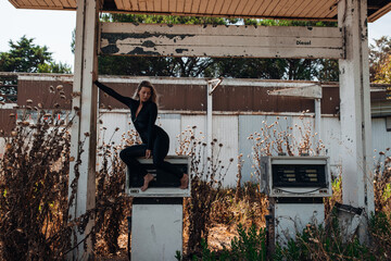 A stylish blonde woman in a black bodysuit poses barefoot on top of an old, weathered gas pump at...