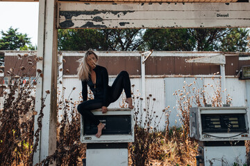 A stylish blonde woman in a black bodysuit poses barefoot on top of an old, weathered gas pump at...