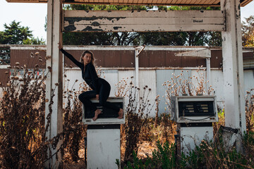 A stylish blonde woman in a black bodysuit poses barefoot on top of an old, weathered gas pump at...