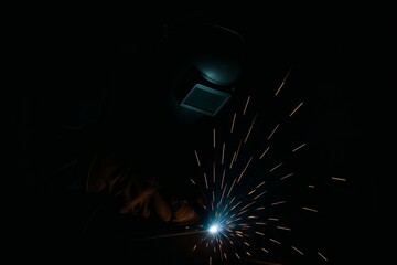 Close-up of a welder working at night in a dark industrial workshop, with bright sparks and a glowing welding arc.