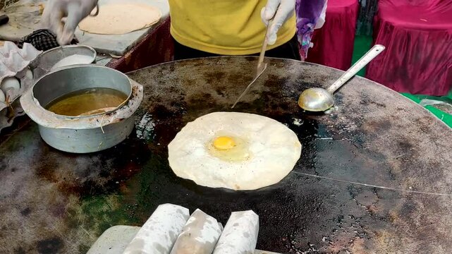 A vendor prepares a delicious egg roll by cracking a fresh egg onto a sizzling paratha on a hot iron tawa. Nearby, trays of neatly wrapped egg rolls, bursting with flavour, await serving. 