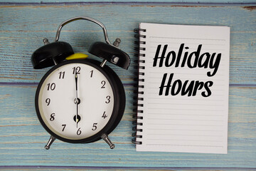 Holiday Hours text written on notepad with alarm clock on wooden background